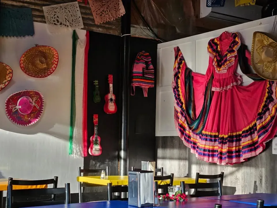 Salle à manger : guirlandes de papel picado, sombreros brodés et robe folklorique au mur, tables jaunes.