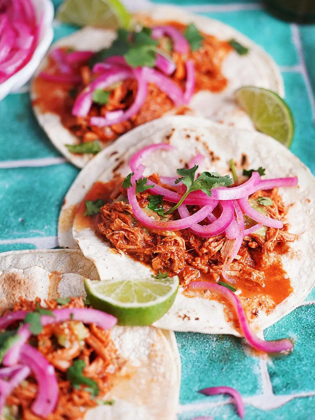 Deux tacos garnis de viande effilochée, d’oignons rouges marinés et d’un quartier de lime, posés sur une table turquoise.