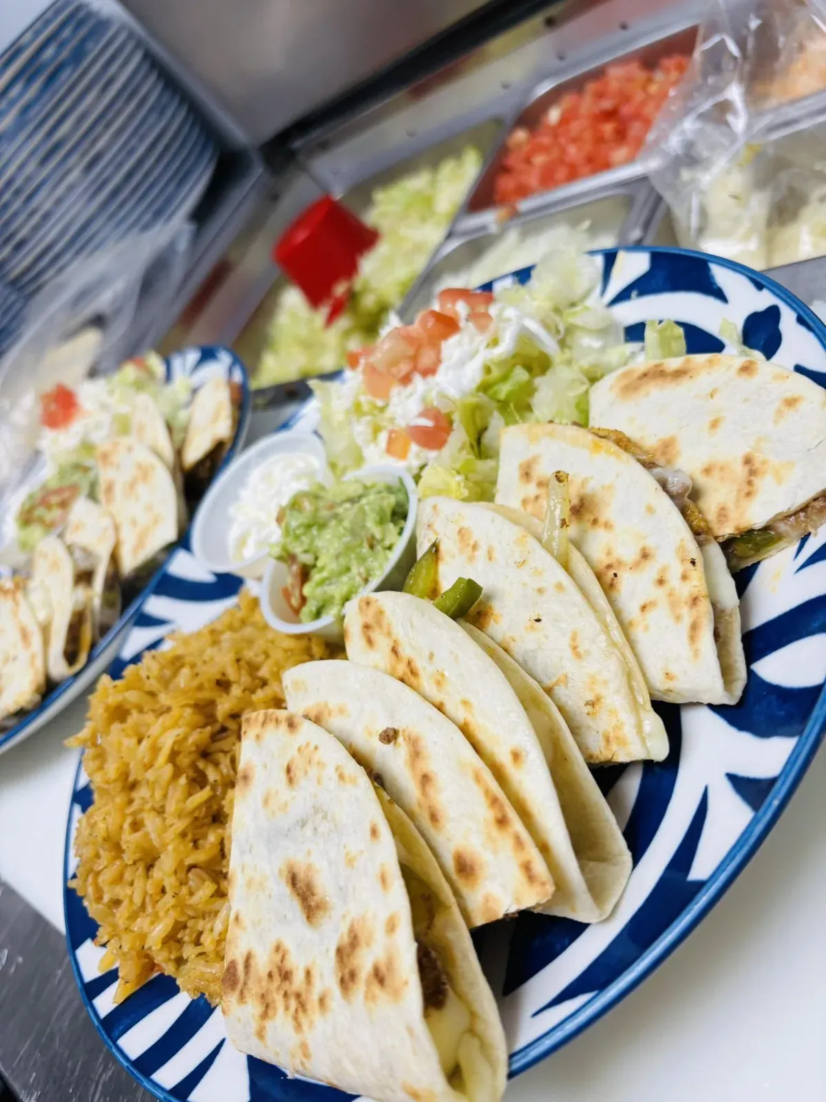 Assiette de talavera bleue et blanche remplie de riz jaune, de tacos souples pliés, d’une coupe de guacamole, de crème sure, de laitue et de tomate coupée.